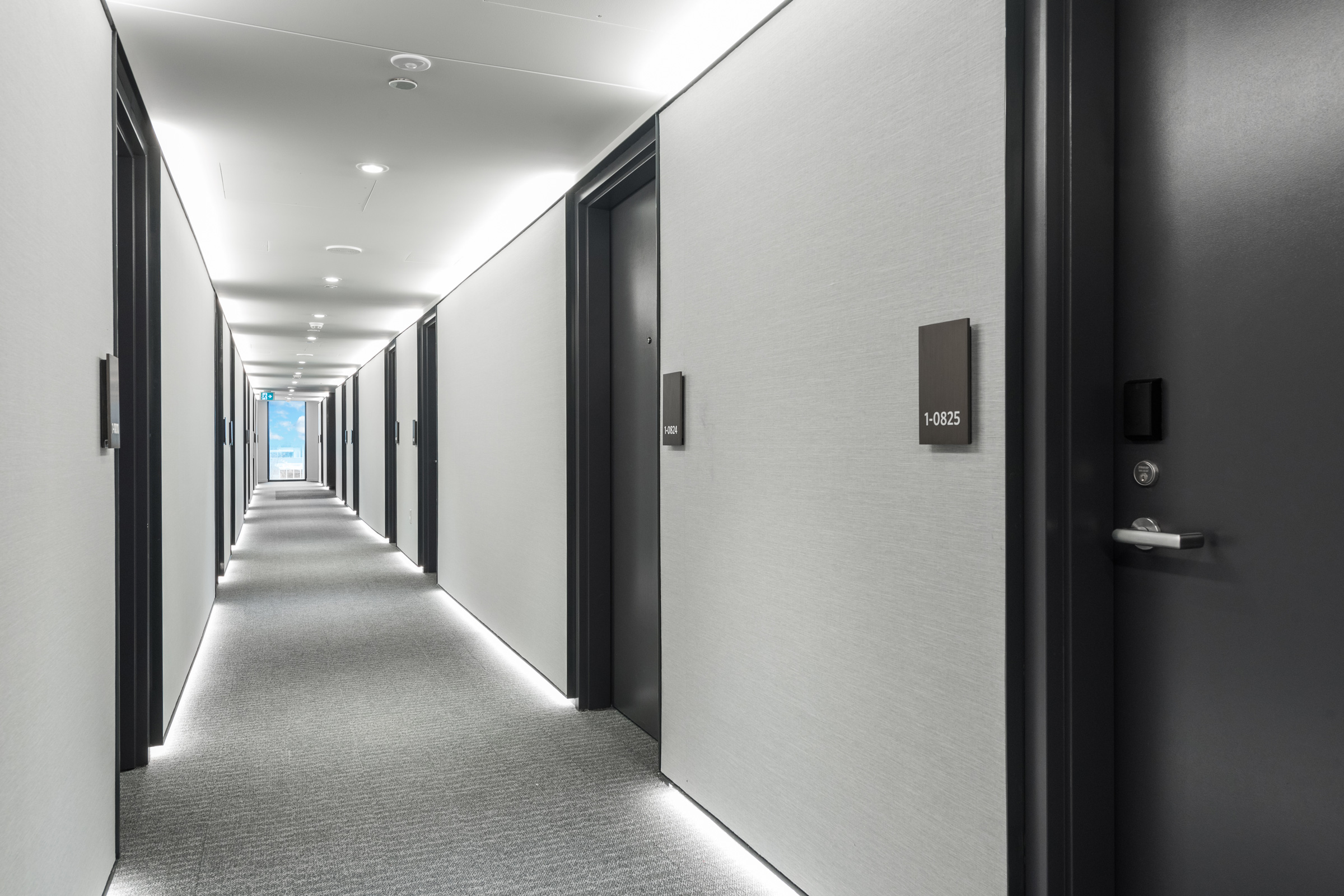 Photo of a beatifully-lit hallway with grey walls, black doors and room number plates, and a full-height window at the end opening onto blue sky at q̓əlɬaləməcən leləm̓