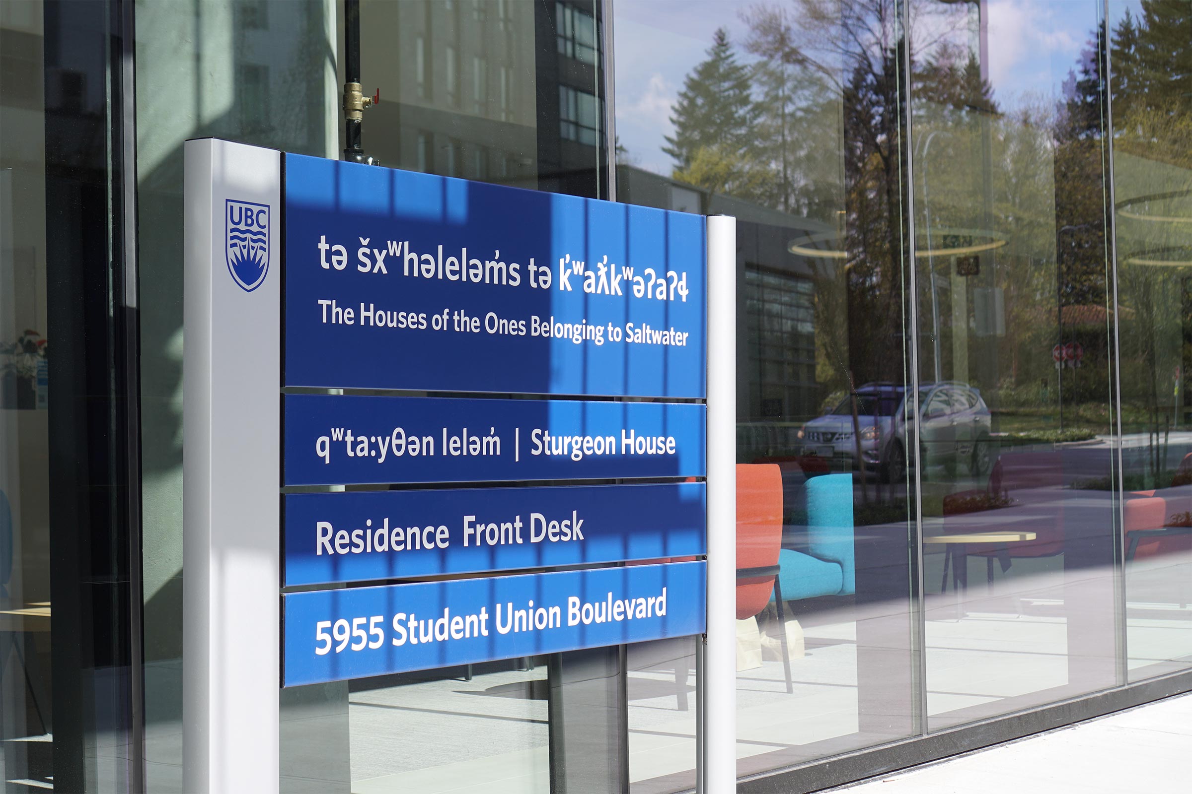 Photo of UBC sign in front of glass windows. Blue and grey UBC building sign with white text that reads tə šxʷhəleləm̓s tə k̓ʷaƛ̓kʷəʔaʔɬ (The Houses of the Ones Belonging to Saltwater", qʷta:yθən leləm̓ (Sturgeon House), Residence Front Desk, 5955 Student Union Boulevard.