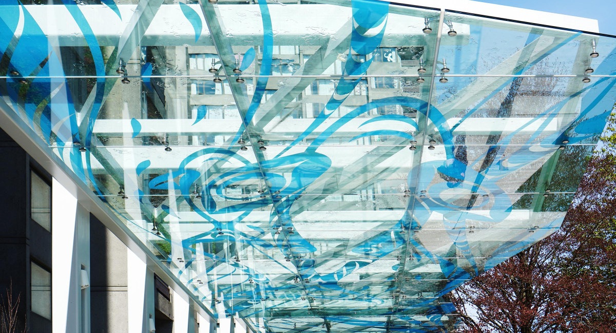 Photo of the glass walkway canopy at tə šxʷhəleləm̓s tə k̓ʷaƛ̓kʷəʔaʔɬ and Walter Gage featuring blue artwork of marine animals by Musqueam artist Kelly Cannell