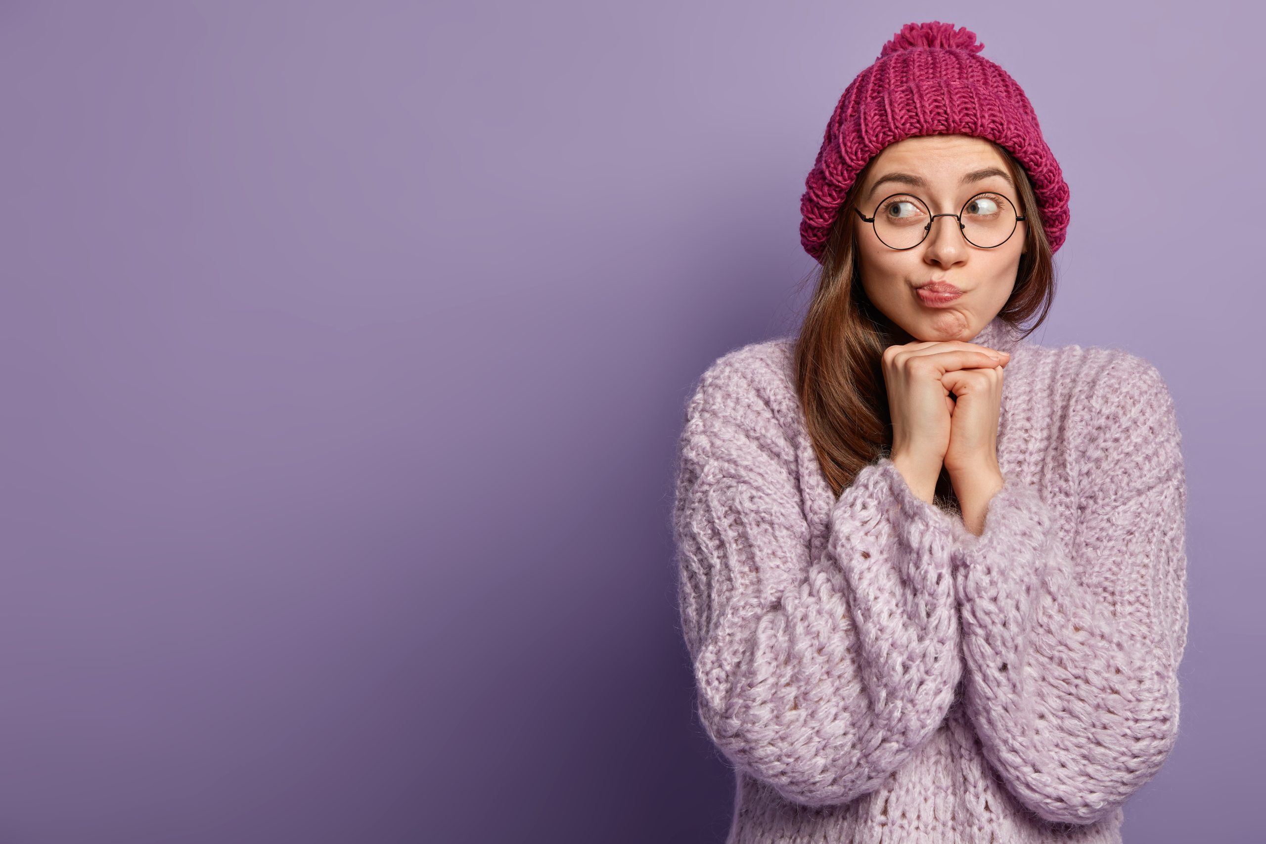 Image of young woman has gaze aside, hands under chin, wears hat and knitted sweater, wears transparent glasses, isolated over purple background